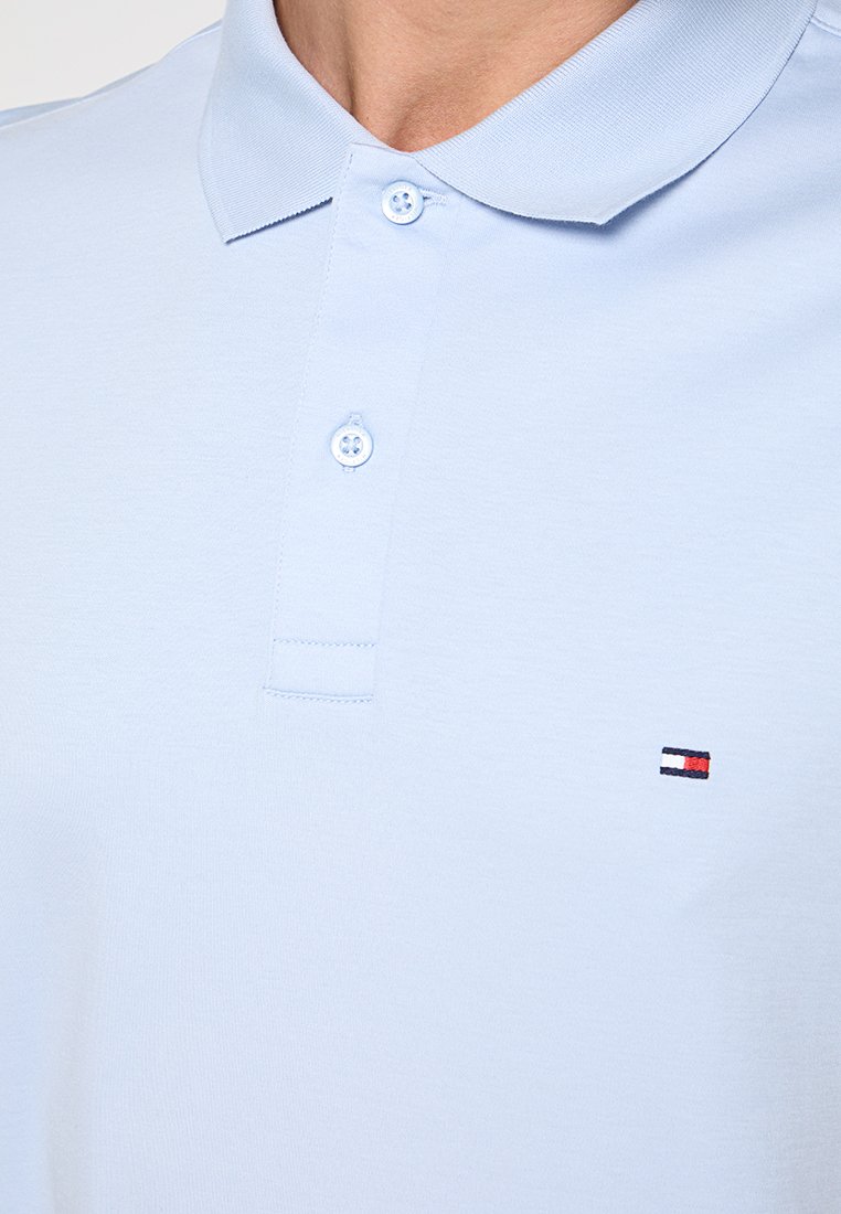 Světle modrá polo košile s dvěma zapnutými knoflíky a malým obdélníkovým logem v červené, bílé a námořnické modři na levé hrudi.