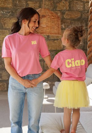 Zwei Personen tragen passende pinke T-Shirts mit gelbem Text. Das T-Shirt des Erwachsenen sagt "Hi", während das des Kindes "Ciao" sagt. Das Kind trägt einen gelben Tüllrock.