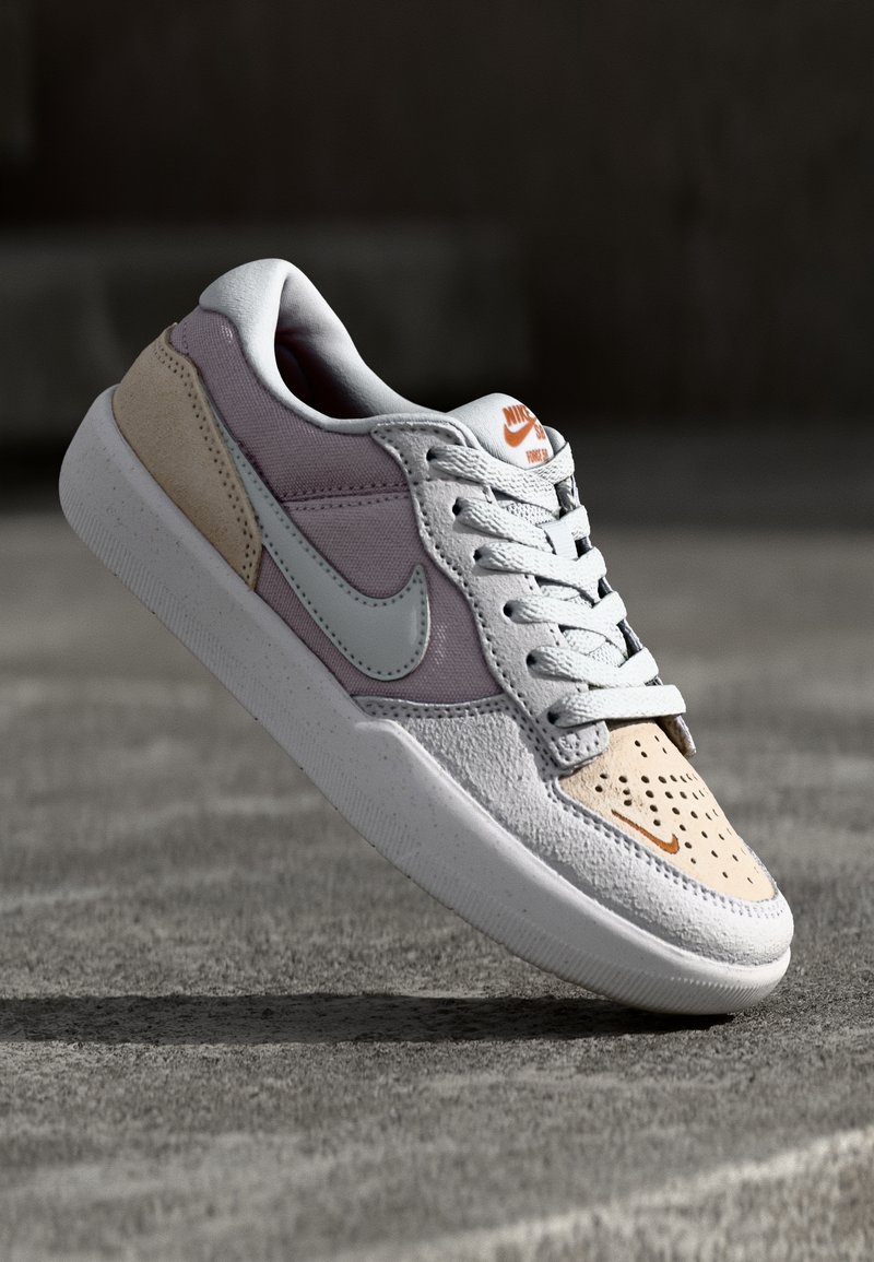 Sneaker Nike a basso profilo con pannelli bianchi, beige e viola chiaro, lacci bianchi e logo Nike arancione, mostrata in volo sopra una superficie in cemento.