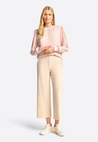 Blouse rose clair avec encolure froncée, manches longues ; pantalon large beige ; baskets blanches avec détails légers.