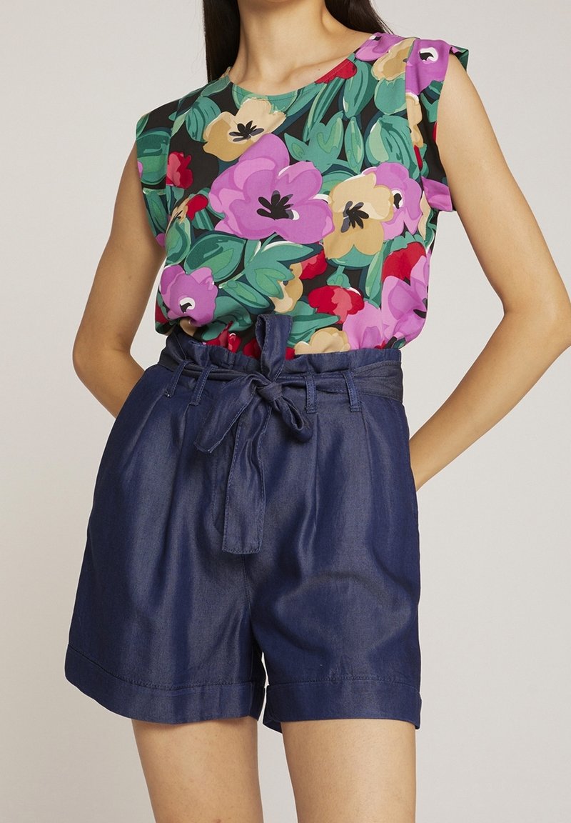 Femme portant un haut sans manches à fleurs aux motifs violets, rouges et beiges, associé à un short taille haute bleu marine noué à la taille.