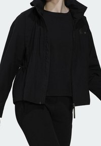Personne portant une veste zippée noire avec un col haut, sur une chemise noire et un pantalon noir, sur un fond clair uni.