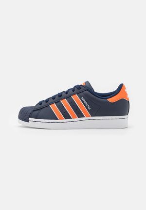 Scarpa Adidas Superstar blu navy con strisce arancioni, linguetta sul tallone e suola bianca, mostrata di profilo su sfondo bianco.