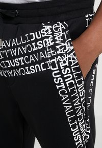 Black sweatpants featuring a textured fabric, an elastic waistband, and repeated white text saying "JUST CAVALLI" along the side.