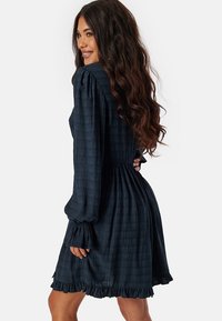 Robe bleu foncé avec un motif texturé, manches longues bouffantes, ourlet volanté et taille cintrée, présentant une silhouette fluide.