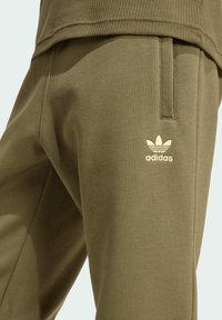 Olive green cotton sweatpants featuring a smooth texture, side pockets, and a cream Adidas logo embroidered on the thigh.