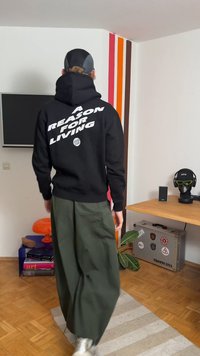 Juodas džemperis su gobtuvu, ant nugaros ryškiu baltu šriftu parašyta "A REASON FOR LIVING", derinamas su laisvomis žaliomis kelnėmis ir sidabriniais sportbačiais.