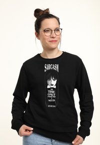Kobieta w okularach i z włosami upiętymi w kok, ubrana w czarną bluzę z grafiką Wednesday Addams i napisem "Sarkazm to jedyny przyjaciel, którego potrzebuję."