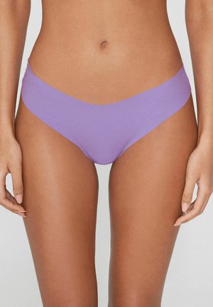 Mujer con braguitas de bikini lila sin costuras, de pie con los brazos relajados a los lados, contra un fondo liso y claro.