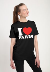 Žena s dlouhými vlasy v culíku, oblečen�á v černém tričku s nápisem "I [červené srdce] PARÍŽ," stojící proti jednoduchému bílému pozadí.