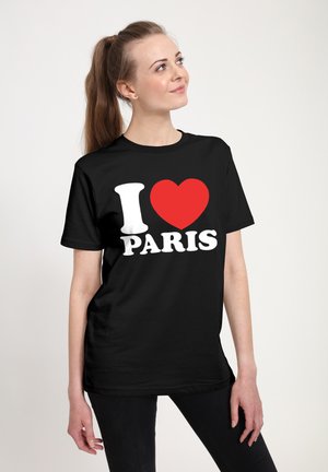 Žena s dlouhými vlasy v culíku, oblečená v černém tričku s nápisem "I [červené srdce] PARÍŽ," stojící proti jednoduchému bílému pozadí.