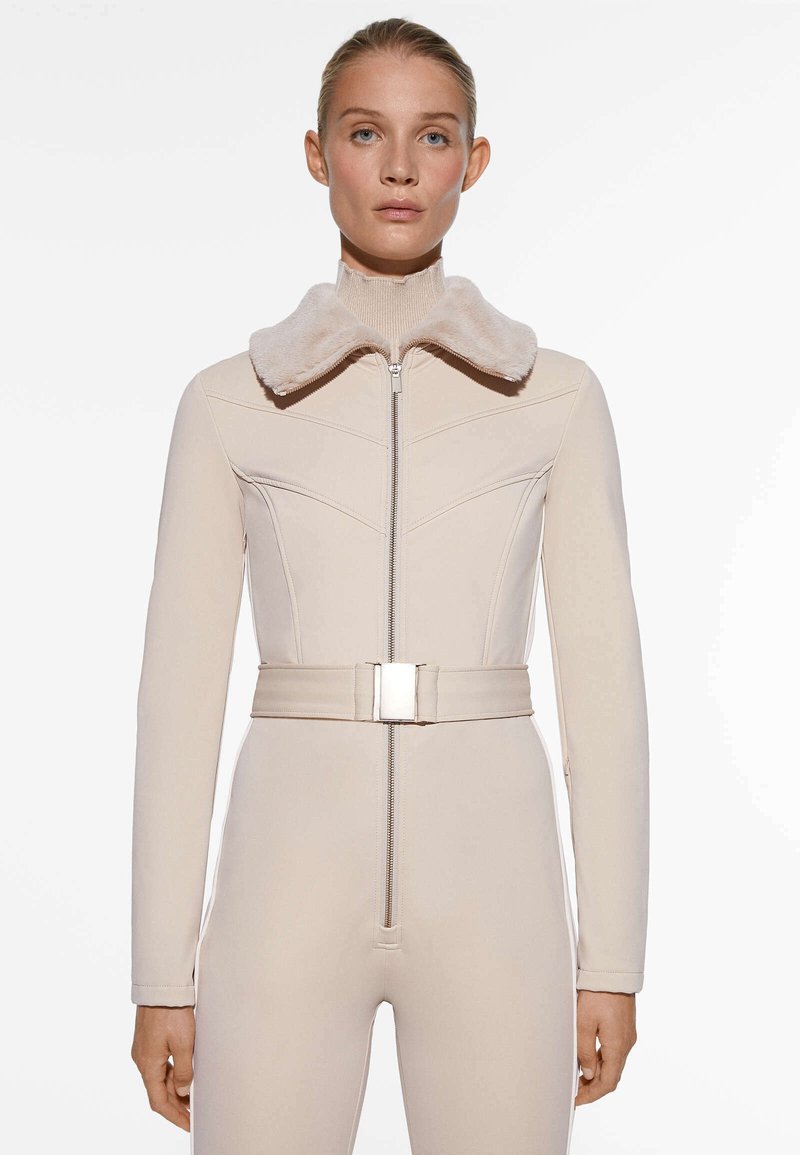 Mono beige con cuello de peluche, silueta ajustada, mangas largas y detalle de cinturón metálico. Textura suave y cierre frontal con cremallera.