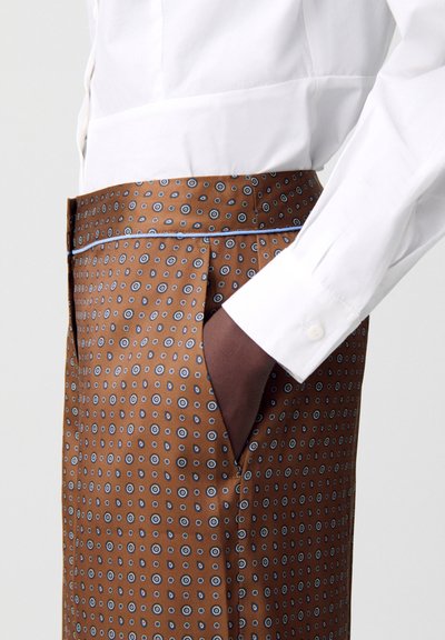 Pantalones marrones con un patrón y motivos circulares azules, con bolsillos laterales y una textura suave, combinados con una camisa blanca ajustada.
