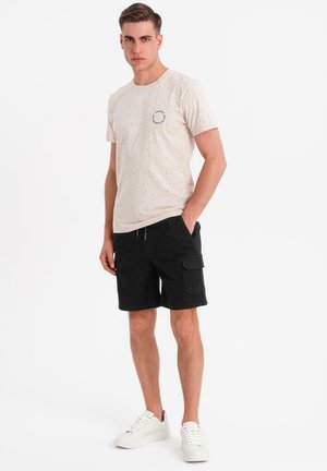 Camiseta de manga corta rosa claro con un patrón moteado y un acento gráfico redondo, combinada con pantalones cortos cargo negros y zapatillas blancas.