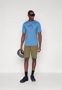 Μπλε κοντομάνικη μπλούζα με σκούρο γραφικό λογότυπο, χακί σορτς, μαύρα αθλητικά παπούτσια και oversized γυαλιά ηλίου. Κρατάει ένα μαύρο κράνος ποδηλάτου.