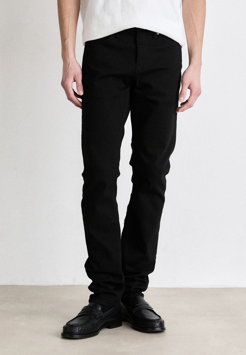 Fekete, slim fit farmernadrág, öt zsebes szabással és diszkrét varrási részletekkel, fekete bőr loafers-zsal párosítva.