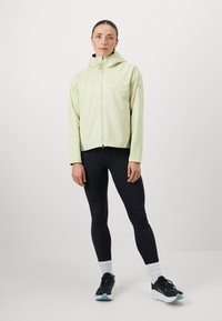 Chaqueta ligera de color amarillo claro con cremallera frontal, capucha integrada y paneles laterales de malla, combinada con leggings negros y zapatillas deportivas.