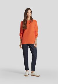 Oranger Strickpullover mit lockerer Passform und gerippten Bündchen, kombiniert mit dunkelblauen Jeans und weißen Sneakers mit hellbraunen Sohlen.