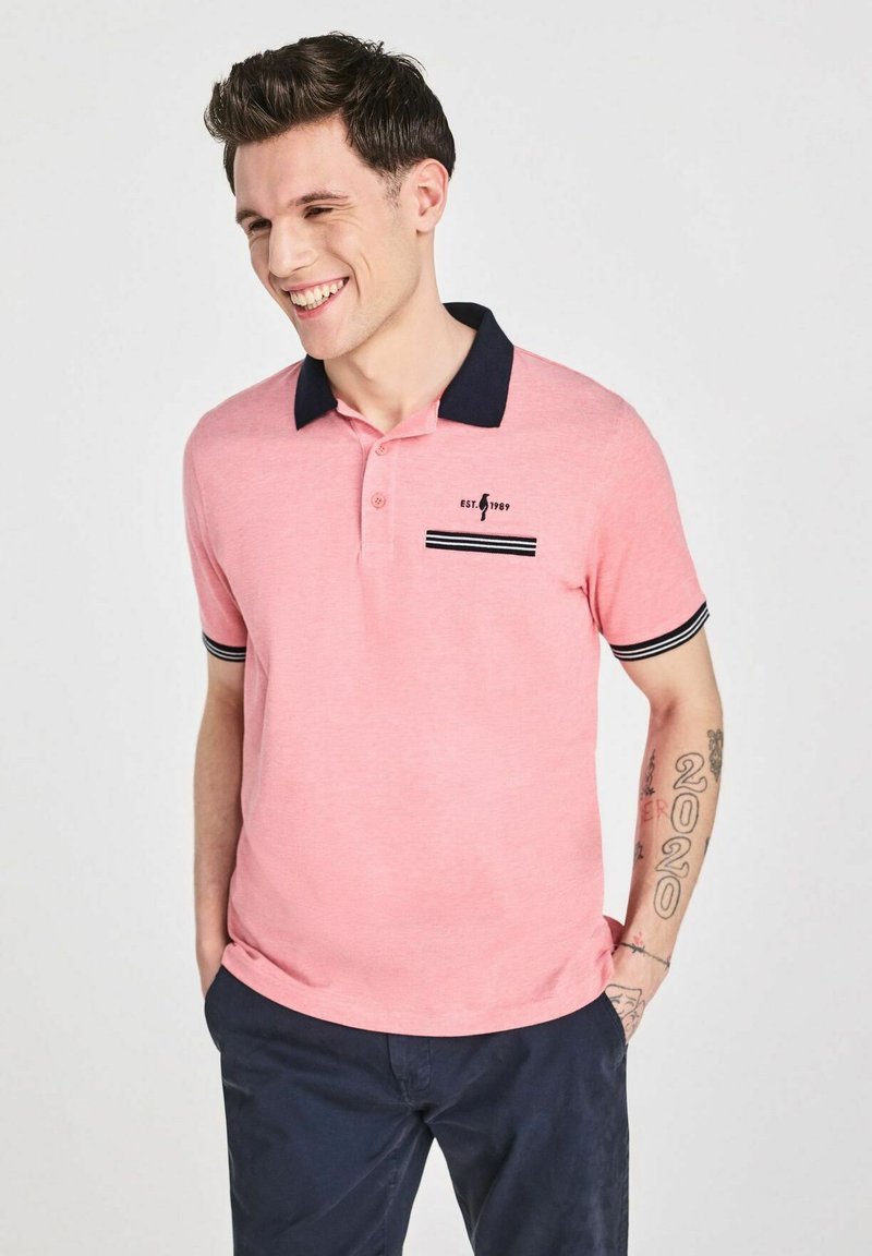 Różowa koszulka polo z granatowym kołnierzykiem i akcentami na rękawach, z małą kieszonką w paski oraz nadrukowanym logo na piersi.