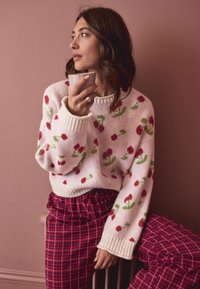 Maglione lavorato a maglia con sfondo rosa, caratterizzato da motivi floreali rossi e foglie verdi. Abbinato a pantaloni scozzesi rossi. Tene in mano una tazza.