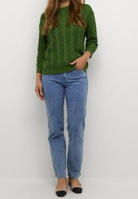 Grüner Kabelstrickpullover mit langen Ärmeln, kombiniert mit hellblauen, geraden Jeans und beigen Ballerinas mit schwarzen Zehenkappen.