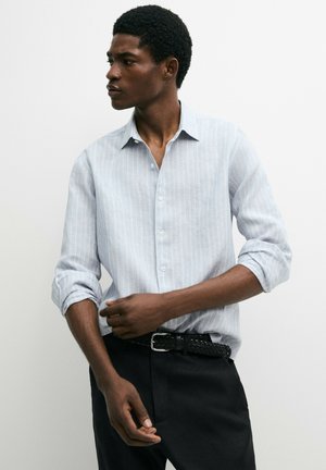 Homme ajustant le poignet d'une chemise rayée bleu clair, portant un pantalon noir et une ceinture noire tressée, regardant sur le côté sur un fond blanc uni.