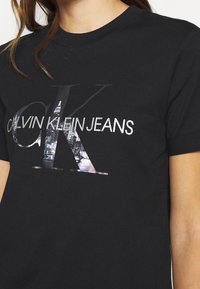 Black cotton t-shirt featuring a prominent graphic in silver and grey depicting "CALVIN KLEIN JEANS" with a cityscape within the letters.