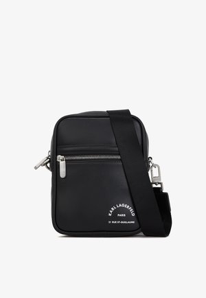 Mala de ombro em pele preta com ferragens prateadas, um compartimento com zíper, uma superfície texturizada e uma marcação branca em destaque na parte frontal.