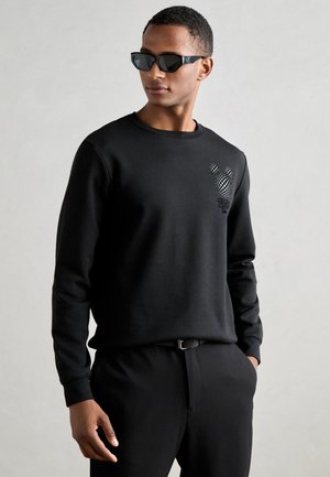 Sudadera negra con cuello redondo, puños acanalados y un diseño de Mickey Mouse a cuadros en el pecho. Combinada con pantalones negros y gafas de sol.