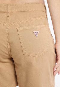 Pantaloni corti beige in cotone con finitura texture. Presentano una tasca posteriore con una patch triangolare del logo Guess.