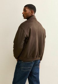 Bruine suède bomberjack met een geribbelde kraag, elastische tailleband en contrasterende stiksels, gedragen over blauwe spijkerbroek.