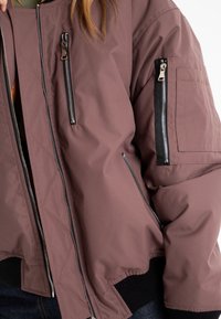 Brun quiltad bomberjacka med flera dragkedjefickor, ribbade ärmslut och en slät yta. Har kontrasterande svarta dragkedjedetaljer.