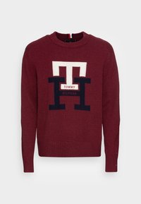 Pull tricoté bordeaux avec col rond, manches longues et un grand logo contrastant au centre avec des éléments blancs et marins.