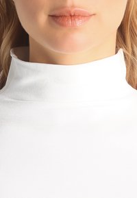 Maglione bianco a collo alto con texture a coste e colletto alto e aderente. Il tessuto ha un aspetto liscio e leggero.