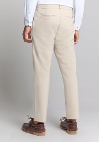 Beige broeken van een gladde stof, met twee achterzakken met paspel en een recht model. Draagt met bruine veterschoenen.