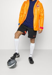 Klart orange vattentät jacka med blå och rosa detaljer, marinblå polo, svarta shorts med röda detaljer, vita fotbollsstrumpor och svarta fotbollsskor.