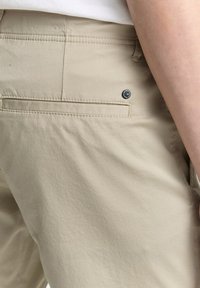 Beige Hose mit glatter Textur, ausgestattet mit einer Gesäßtasche, dezenten Stichdetails und einem runden schwarzen Hardware-Akzent.