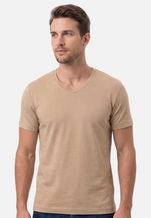 Junger Mann mit hellbraunem Haar, der ein beigefarbenes T-Shirt mit V-Ausschnitt und kurzen Ärmeln trägt, leicht nach links gewandt vor einem einfarbigen weißen Hintergrund.