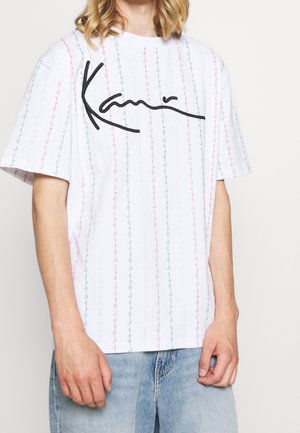 Personne portant un t-shirt blanc oversize avec des rayures verticales multicolores et un grand logo noir en cursive sur la poitrine.