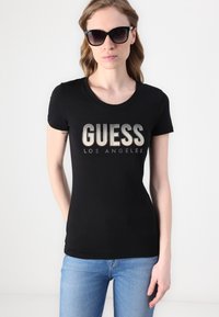 Melna īsā piedurknē T-kreklu ar metālisku "GUESS LOS ANGELES" tekstu, izrotātu ar kristāliem. Valkāts ar zilo džinsu biksēm.