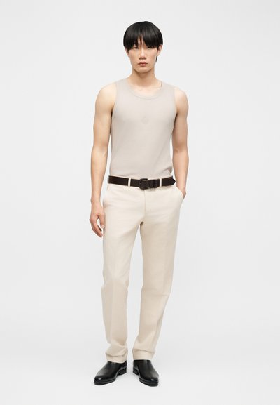 Άνδρας με μαύρα μαλλιά που φοράει μπεζ αμάνικη μπλούζα, κρεμ παντελόνι με μαύρη ζώνη και μαύρα παπούτσια, στέκεται με το ένα χέρι στην τσέπη.