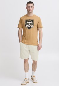 Brown cotton t-shirt with a graphic of a panda wearing sunglasses. Paired with light beige shorts and beige athletic shoes with stripes.