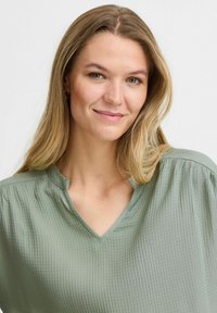 Blusa di un verde chiaro, con texture, scollo a V e maniche corte. Presenta pieghe sottili lungo le spalle per un tocco di dettaglio in più.