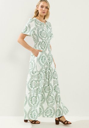 Frau in einem weißen Maxikleid mit grünen, kreisförmigen Blumenmustern und kurzen Ärmeln, kombiniert mit braunen Sandalen mit Absatz, steht vor einer schlichten Wand.