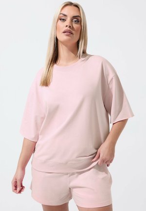 Blondynka w luźnej, jasnoróżowej bluzce z krótkim rękawem i pasujących szortach, stojąca na tle jasnego, jednolitego tła.