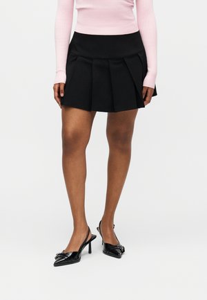 Kvinne iført svart plissert miniskjørt, rosa langermet ribbestrikket topp og svarte spisse høyhælte slingback-sko står på hvit bakgrunn.