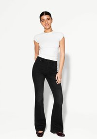 Weißes, eng anliegendes Kurzarmoberteil mit Rundhalsausschnitt, kombiniert mit hochtaillierten, schwarzen Schlagjeans. Das Outfit wird durch dunkel gefärbte Schuhe ergänzt.