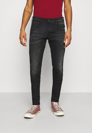 Mand iført falmede sorte skinny jeans, røde høje sneakers og en rød skjorte, der står foran en ensfarvet baggrund.