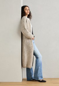 Long beige knit cardigan with a relaxed fit, paired with a white top and flared blue jeans. Black flats complete the outfit.