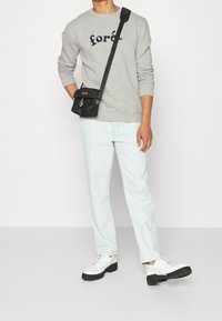 Persoon in een grijze sweatshirt met de tekst "forét", lichtblauwe jeans, witte schoenen en een zwarte schoudertas met oranje details.